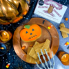 Billington's Gingerbread and an Original Biscuit Bakers pumpkin biscuit displayed on a copper serving platter in a Halloween-themed setting, surrounded by candles, cobwebs, candy corn, skeleton hand decorations and seasonal spooky props.