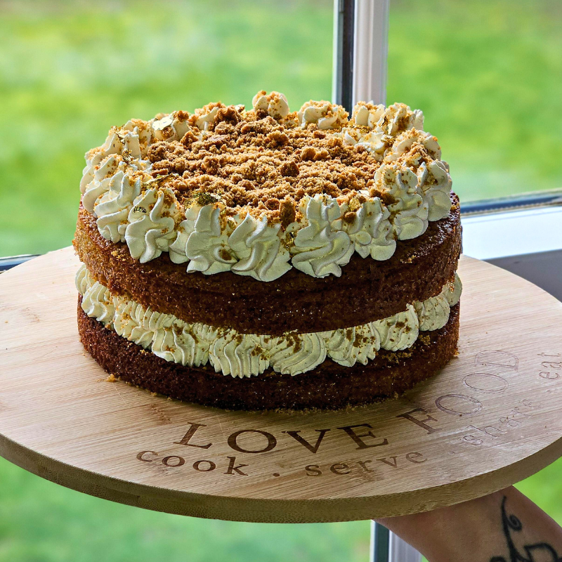 Handmade two‑layer sponge cake with generous piped cream filling and topping, finished with crunchy biscuit crumbs on a wooden engraved serving board.