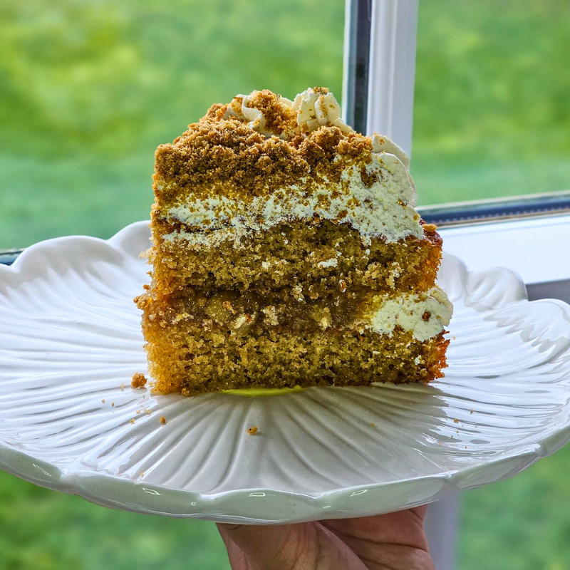 Slice of Billington’s Gingerbread Apple and Ginger Sandwich Sponge with creamy filling and biscuit crumb topping on a white decorative plate.