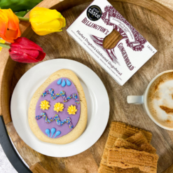 Original Biscuit Bakers Easter egg sugar cookie with colourful icing, served with Billington’s Gingerbread fingers, tulips, and a warm drink on a rustic wooden tray.