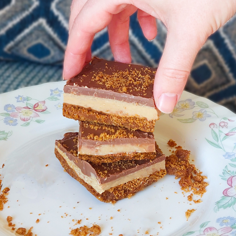 Hand lifting a slice of millionaire’s gingerbread with a spiced gingerbread base, fudge layer and milk chocolate topping, finished with gingerbread crumbs.