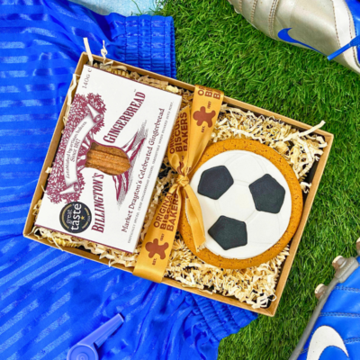 Football‑shaped iced gingerbread biscuit and Billington’s gingerbread presented in a gift box, styled with blue football boots and kit on artificial grass.