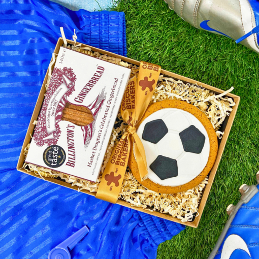 Football‑shaped iced gingerbread biscuit and Billington’s gingerbread presented in a gift box, styled with blue football boots and kit on artificial grass.