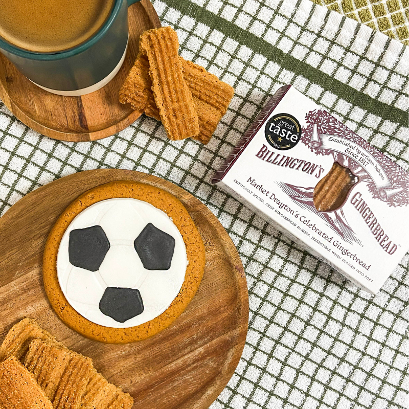 Football‑shaped iced gingerbread biscuit on a wooden board, styled with Billington’s gingerbread, loose gingerbread biscuits, and a mug on a checked kitchen cloth.