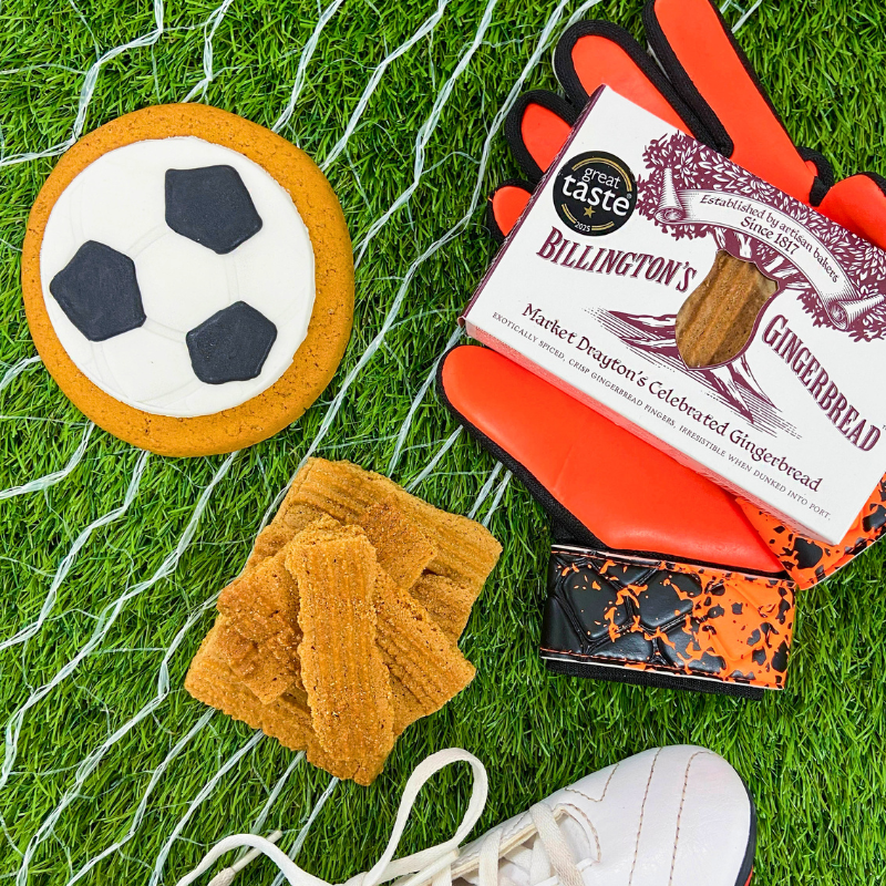 Football‑shaped iced gingerbread biscuit displayed with Billington’s gingerbread, goalkeeper gloves, and football boots on artificial grass with goal net.